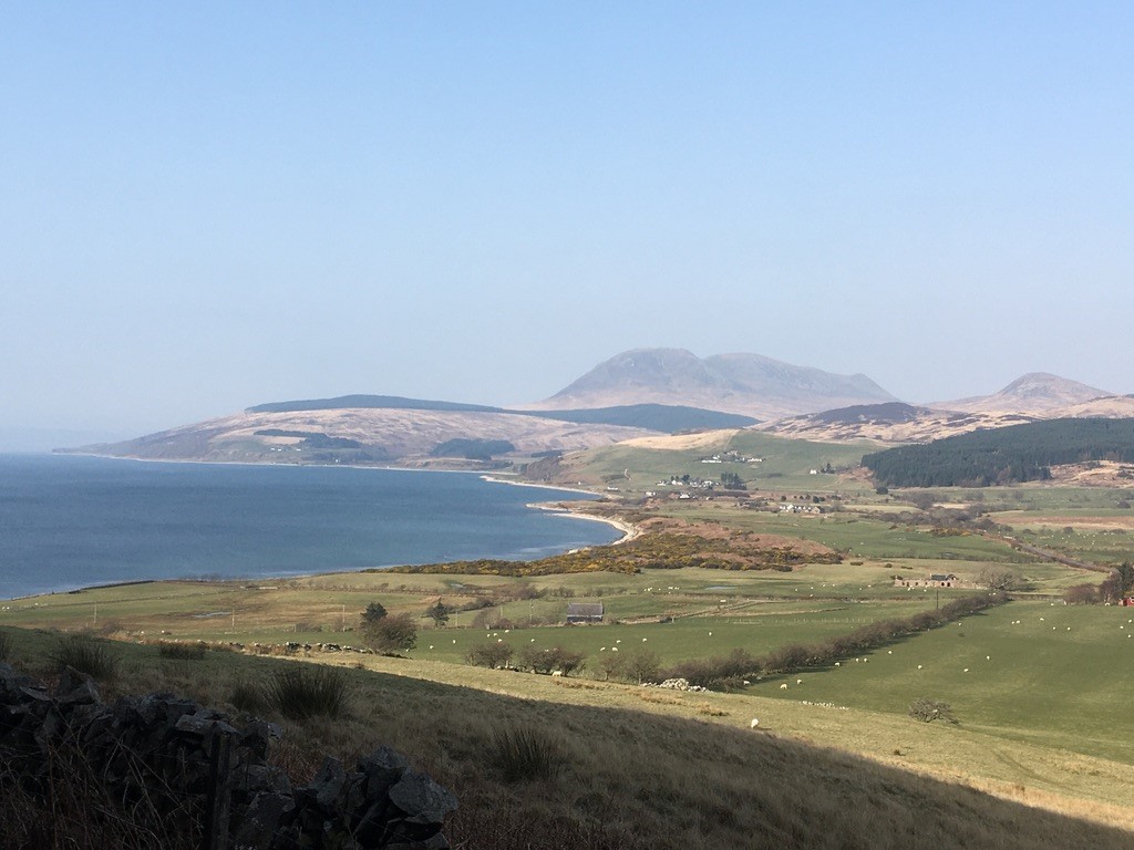 The photograph shows the landscape of Machrie Bay from the south. There is a clear blue sky, reflected in the sea to the left. The land to the east shows grazing land, forestry and mountains in the distance.